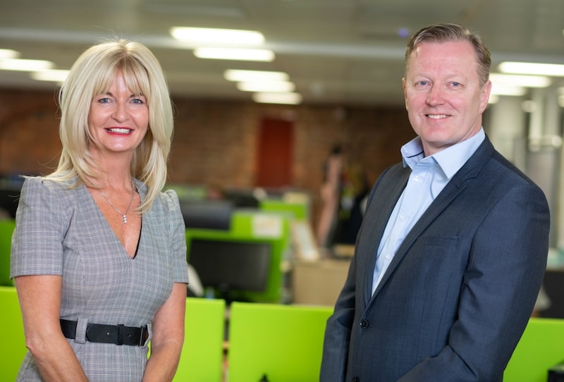 Two people in business attire smiling in a modern office setting with bright lighting and green chairs in the background.